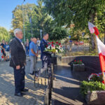 Na obrazku grupy mężczyzn, z których są ubrani w formalne stroje, umieszczając je na miejscu przy pomniku polskimi flagami. W tle znajdują się strażacy oraz osoby podlegające.