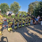 Na obrazie znajduje się grupa podlegająca w mundurach, ustalona na baczność przed tłumem obserwatorów, zarówno dzieci jak i dorośli. Cała scena ma miejsce podczas słonecznego dnia.