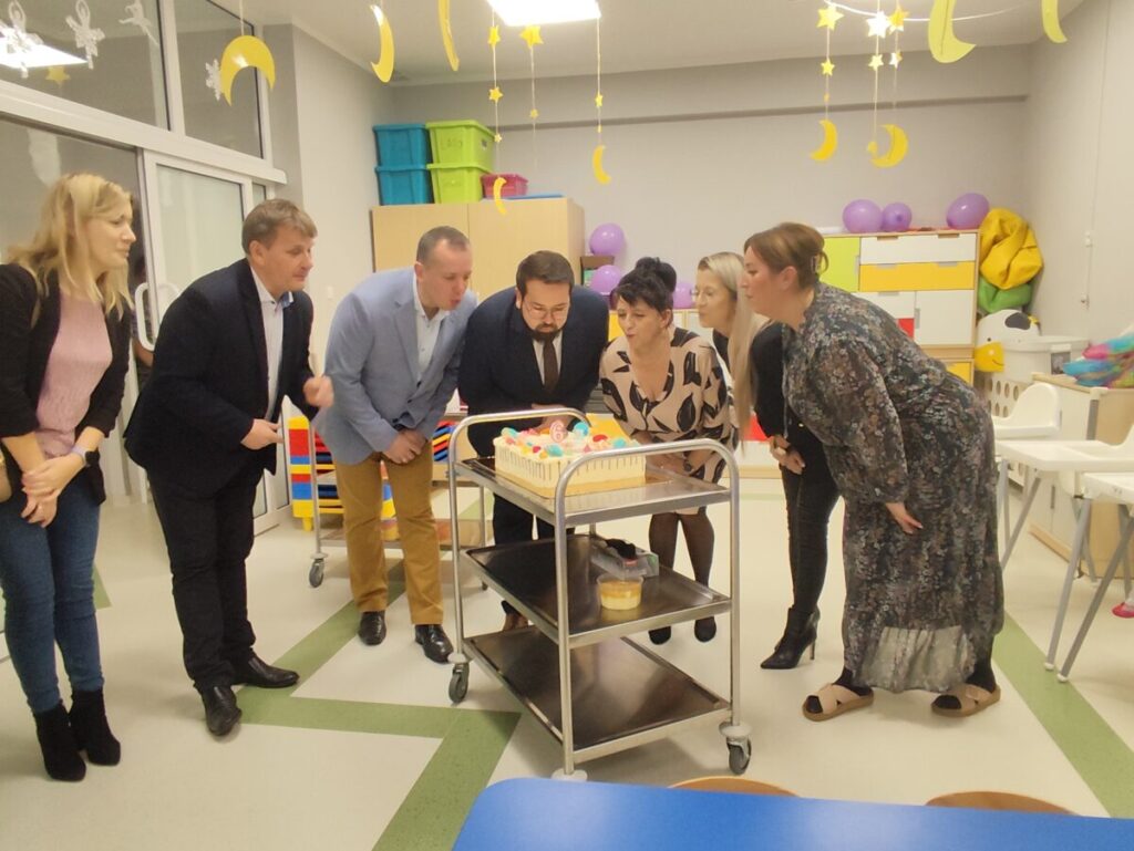 Na obrazku jest grupa osób zgromadzona wokół tortu umieszczonego na wózku w jasnym, kolorowo udekorowanym pomieszczeniu. Z sufitu zwisają dekoracje w formie gwiazd i księżyców, tworząc świąteczną atmosferę.