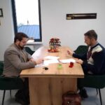 Dwóch mężczyzn siedzi przy drewnianym ukradł w i podpisuje dokumenty. W środku znajduje się poinsecja i misa z owocami, a w tle są widoczne duże okno oraz dekoracyjne ściany.