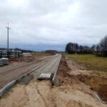 Na obrazie widać nieutwardzoną drogę w trakcie budowy, z hałdami materiałów budowlanych po jednej stronie. Droga otoczona jest przez bezlistne drzewo oraz pole, a nad krajobrazem pochmurne niebo.