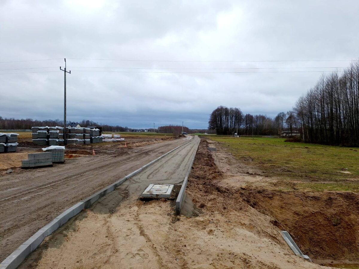 Na obrazie widać nieutwardzoną drogę w trakcie budowy, z hałdami materiałów budowlanych po jednej stronie. Droga otoczona jest przez bezlistne drzewo oraz pole, a nad krajobrazem pochmurne niebo.