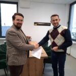 Na obrazku widoczni są dwaj mężczyźni wymieniający się dokumentem podczas uścisków dłoni w biurze. Jeden z nich ma na sobie marynarkę, drugi sweter; w tle znajduje się stół z kwiatami.