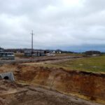 Na zewnątrz znajduje się miejsce budowy z ułożonych stosami betonowych bloków na otwartym terenie, a niebo jest zachmurzone. W tle widać słupy energetyczne oraz kilka domów.