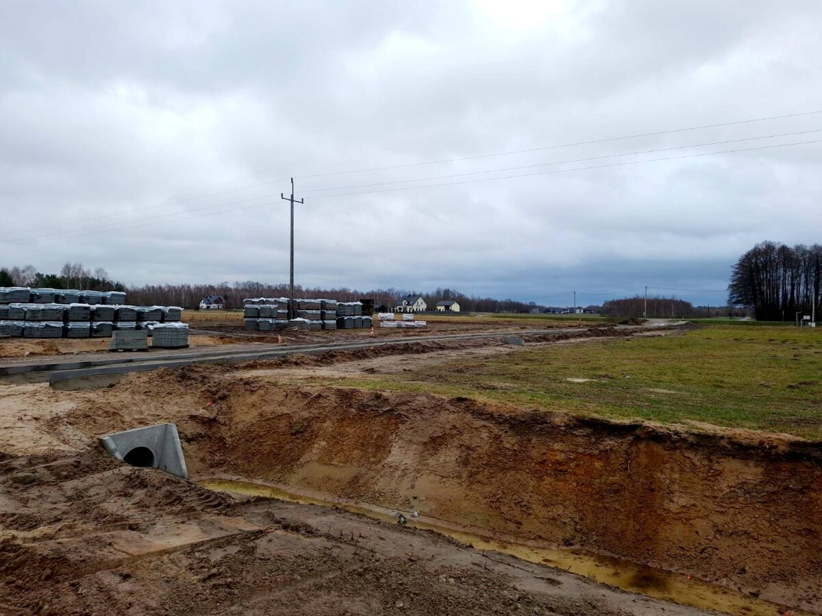Na zewnątrz znajduje się miejsce budowy z ułożonych stosami betonowych bloków na otwartym terenie, a niebo jest zachmurzone. W tle widać słupy energetyczne oraz kilka domów.