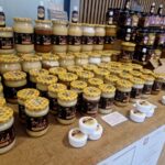 Na miejscu znajduje się kilka opakowań miodu o różnych rozmiarach i etykietach, z przodu, są małe, białe pojemniki. Delikatna substancja kolorowa i zachęcająca do prezentowania produktów pszczelarskich.