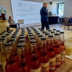 Na pierwszym planie znajdują się butelki i pojemniki na stole. W tle widać osobę przemawiającą na terenie obok ekranu prezentacyjnego, a po prawej stronie siedziby.