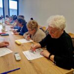 Starsze kobiety siedzą przy stole i ustalają się na wypełnianiu formularzy za pomocą długopisów. Na stole znajdują się różne przedmioty, takie jak pilot do telewizora i piórniki, a w jasnym pomieszczeniu widać ludzi w tle.