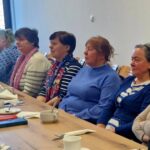 Na zdjęciu jest grupa osób siedząca przy zamkniętym stole, zawarta w konsultacjach lub regulaminie. Na stole znajdują się filiżanki i talerze, co jest charakterystyczne dla napojów i przekąsek.