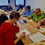 Starsza grupa osób siedzi przy drewnianym, dobrze oświetlonym pomieszczeniu, skupiając się na pisaniu i czytaniu dokumentów. Trzymają się w dłoniach długopisy, podczas gdy inni koncentrują się na zawartości przed sobą.