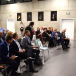 Na widocznym jest mówca przemawiający do uczestnika zgromadzonej w nowoczesnej sali konferencyjnej. ubrani, wszyscy mężczyźni, jak i kobiety ubrani w biznesowe stroje, z uwagą słuchają, z nich robią notatki.