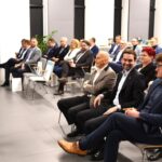 Na obrazku widać mówcę ubranego w kolorze niebieskim, który jest przewodniczącym zgromadzonej w nowoczesnej sali konferencyjnej. ubrani w ubrania biznesowe, słuchają z uwagą.