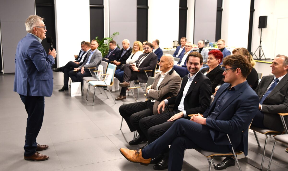 Na obrazku widać mówcę ubranego w kolorze niebieskim, który jest przewodniczącym zgromadzonej w nowoczesnej sali konferencyjnej. ubrani w ubrania biznesowe, słuchają z uwagą.