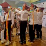 480619460_662747669434091_2503944608230819395_n Na obrazku widoczne są dzieci ubrane w kolorowe stroje i czerwone czapki, które słonecznie tańczą trzymając się za ręce oraz trzymając wstążki. wystąpienie zjawiska na drewnianej instalacji w jasnooświetlonym pomieszczeniu.