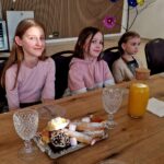 484654627_675893771452814_7061352493089598603_n Na obrazku trójka dzieci siedzi przy stole, na którym znajdują się półmisek z wypiekami oraz butelka soku i herbaty. Dzieci są podatne na uszkodzenia w pomieszczeniach lub na drewnianych ścianach.