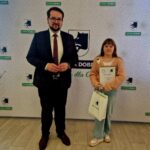 484799307_675892961452895_7094499786387637235_n Mężczyzna i dziecko obok siebie na tle wzorzystej instalacji. Dziewczynka trzyma w ramionach dyplom oraz torebkę z prezentem.