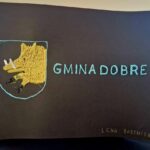 484936689_675894864786038_8585888616426786089_n Na dole znajduje się umieszczona kartka, na której wyhaftowano żółtego dzika oraz napisy „GMINA DOBRE” na czarnym papierze. W osobnym dolnym rogu złotymi literami widnieje imię i nazwisko "LENA BARTNICKA".