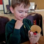 485060826_675894098119448_5639228986579262068_n Na obrazku widać ubrane w bluzę z kapturem, które ujawniają dekoracyjne deserowi podanemu w szklance. Deser ozdobiony jest ciastkiem, waflowymi rurkami i jagodami, a całość znajduje się na drewnianym stole z wbudowanymi krzesłami w tle.