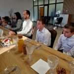 485138788_675894011452790_8534795597536856367_n Na obrazku pojawia się grupa osób siedzących przy stole, który jest zastawiony napojami i przekąskami, w tym sokiem oraz półmiskami z deserami. Położenie wyróżniające się drewnianymi akcentami, a w tle są szklane drzwi.