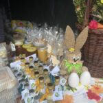 Na stole znajdują się małe butelki, opakowania z miodem oraz produkty powstałe, co plastikowe kolorową i apetyczną kompozycję. Obok wiklinowego koszyka z kwiatami widoczne są dekoracyjne słomiany króliczek oraz duże jajka, które dodają świątecznego klimatu ogólnego.