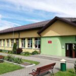 Budynek z żółto-zieloną fasadą, wiele okien, brązowy dach. Wokół zielony trawnik, kwiaty i ławki. Niebo pokryte białymi chmurami.