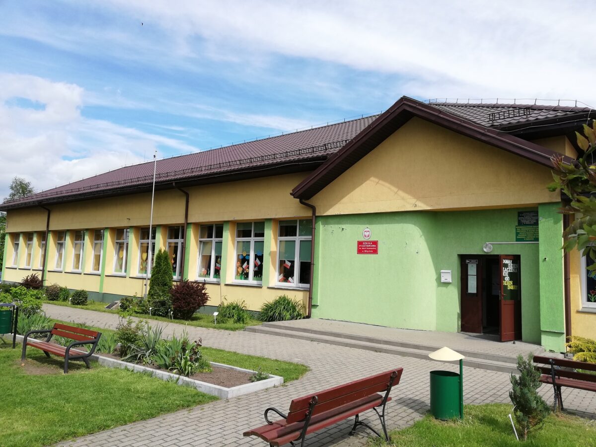 Budynek z żółto-zieloną fasadą, wiele okien, brązowy dach. Wokół zielony trawnik, kwiaty i ławki. Niebo pokryte białymi chmurami.