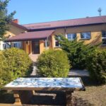 Na zdjęciu widać żółty budynek z brązowym dachem, otoczony zielonymi krzewami. Na pierwszym planie stoi stół z mozaiką. W tle błękitne niebo i drzewa.