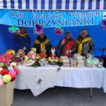 Na zdjęciu cztery osoby stosowane za stołem, które zawierają opis rękodzieła i wypieki. Przedmiot pokazywany jest pod niebieskim namiotem z napisem "Koło Gospodyń Wiejskich Dobrzynianki".