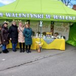 Na obrazku znajduje się pięć osób stojących przed zielonym namiotem udekorowanym o tematyce związanej z Wielkanocą, obok której znajduje się krzesło z wystawionymi przedmiotami. Namiot jest opatrzony banerem z napisem w języku polskim.