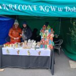 Na obrazku znajdują się dwie osoby, które są za stołem, na których są wystawione herbatyi z przetworami, chleb i rękodzieło. znajdujący się pod zielonym namiotem opatrzonym napisem „ZAKRĘCONE KGW w dobrym”, co wskazuje na prezentację produktów przygotowanych przez Koło Gospodyń Wiejskich w lokalizacji Dobre.