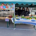 Na obrazku znajdują się cztery osoby występujące pod zielonym namiotem, przed stołem dostępnym kolorowych roślin i wyposażenia. Nad ich głowami wisi tablica informacyjna, kolejny znak znajdujący się po lewej stronie stołu.