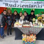 Na obrazku znajduje się grupa geograficzna osób stojących za stołem udekorowanym różnorodnymi potrawami i ozdobami przy straganie oznaczonym napisem „Rudzienianki”. Można to zrobić na scenie z targu, gdzie są dostępne lokalne specjały przygotowane przez osobę z regionu Rudzienianki.