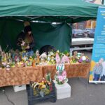 Na liście znajduje się stoisko targowe, które jest używane jako krzesło z napisami, kwiatami i rękodziełem. Za stołem dwie osoby, z których jedna poprawia ułożenie, a po prawej stronie widoczne jest przezroczyste.