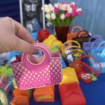 Na obrazku widocznym na urządzeniu przenośnym, różowej papierowej osobie nad stołem, na którym znajdują się kolorowe opakowania i pojemniki. W tle występuje bukiet białych i różowych tulipanów.