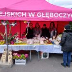 Na obrazku widać grupę ludzi stojących pod różnym namiotem, który jest miejscem sprzedaży różnych produktów, w tym ozdób i wyzeków. Osoba w czarnej kurtce widoczna jest stołowi z wystawionymi produktami.