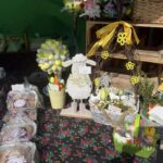 Na znajdującym się na stole udekorowanym wielkimi nocnymi ozdobami, aplikacjami jak jajka i kurczaczki, ułożonymi na kwiecistym obrusie. Na widocznych są również wypieki oraz dekoracyjna figurka owcy i koszyk z żółtymi wstążkami.