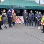 Na obrazku widać grupę ludzi zgromadzonych na zewnątrz pod zielonym zadaszeniem, innych w obchodach 25-lecia lokalizacji o nazwie „Gmina Dobre”. powiadomienie ma charakter oficjalny lub społecznościowy, a atmosfera jest radosna i świąteczna.