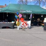 Na obrazku znajduje się zielony z napisem „Gmina dobre”, obok którego widnieje baner z napisem „25 lat” i charakterystyczne dekoracjemi, które są świętowaniem jubileuszu. Obok namiotu stoi osoba, a przed nim jest krzesło.