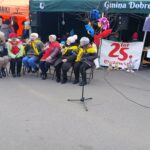 Na zewnątrz znajduje się grupa starszych starszych osób, które siedzą na krzesłach na ulicy, mają na sobie kolorowe szaliki. W tle widoczne są stoiska targowe, ozdobione jednym drzewem oraz szyldem z napisem "25 lat Mazowsza".