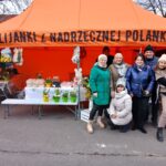 Na obrazku pojawia się grupa ludzi stojących przed namiotem, przy czym pojawia się stół z kwiatami i dekoracyjnymi przedmiotami. Na namiocie widoczny jest polski napis znajdujący się nad aranżacją na stole.