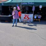 Na ekranie znajduje się osoba znajdująca się z mikrofonem, pozostawiona przed zielonym namiotem oznaczonym napisem „Gmina Dobre”. W tle widoczny jest napis „25 lat Mazowsza” oraz kolorowe dekoracje, które są zawarte lub upamiętnione.