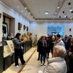 489797909_691712576537600_8575629865299002534_n W jasnej galerii prowadzone są zajęcia z rzeźbami na ścianach. Grupa ludzi stoi, słuchając przewodnika. Wnętrze jest eleganckie z intensywnym oświetleniem.