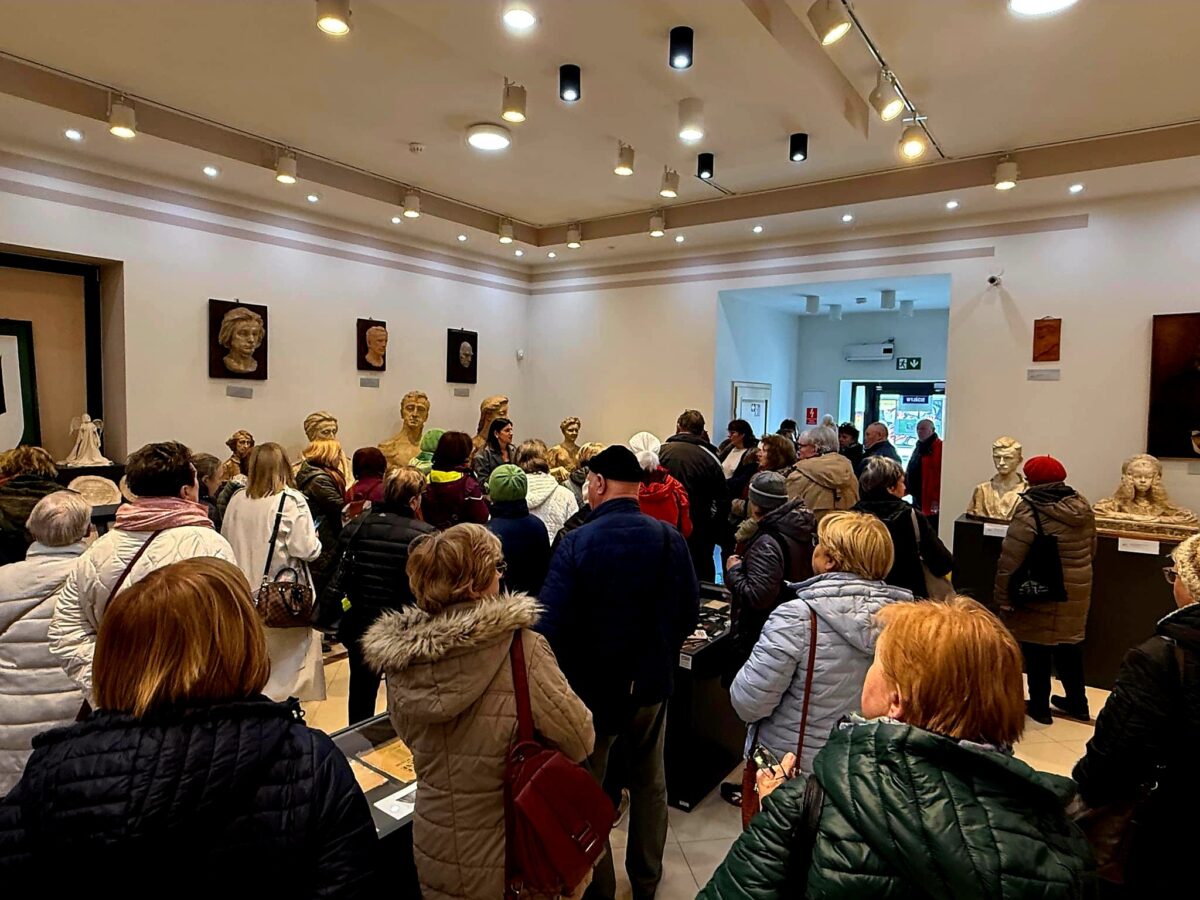 Grupa ludzi ogląda rzeźby w jasnej galerii. Na ścianach wiszą portrety w neutralnych kolorach. Tłum nosi kolorowe kurtki.