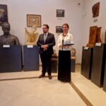 Dwoje ludzi w eleganckich strojach stoi w galerii sztuki. W tle widoczne są rzeźby i malowidła na jasnych ścianach. Podłoga jest wyłożona kafelkami w ciepłych odcieniach beżu.