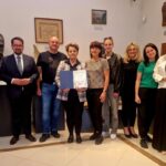 Na obrazku grupa ośmiu osób stoi w muzeum. Tło zdobią rzeźby i obrazy w neutralnych kolorach. Centralna osoba trzyma dyplom.