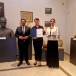 Trzy osoby stoją w muzeum, trzymając dyplom. Tło to popiersia i obrazy na ścianie. Dominują brązowe i szare kolory.