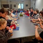 Grupa osób siedzi przy długim stole, zajadając przekąski i pijąc napoje. W tle widać jasne pomieszczenie z oknami. Stół zdobi zielona roślina.