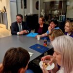 Grupa ludzi siedzi przy szarym stole w jasnym pomieszczeniu. Na stole leżą niebieskie teczki i napoje. W tle szklana witryna z eksponatami.