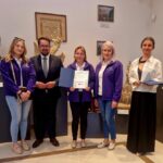 Grupa osób pozuje w muzeum obok rzeźb. Trzy osoby w fioletowych kurtkach trzymają dyplom. W tle obrazy i popiersia na ścianie.