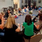 Na zdjęciu grupa ludzi siedzi przy dużym stole w jasnym pomieszczeniu. Stoły ozdobione są sztuczną zielenią i napojami. Ludzie rozmawiają i piją napoje.